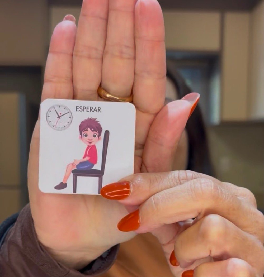 Mão de um adulto segurando o cartão ESPERAR do guia, com o desenho de uma criança sentada numa cadeira e um relógio.